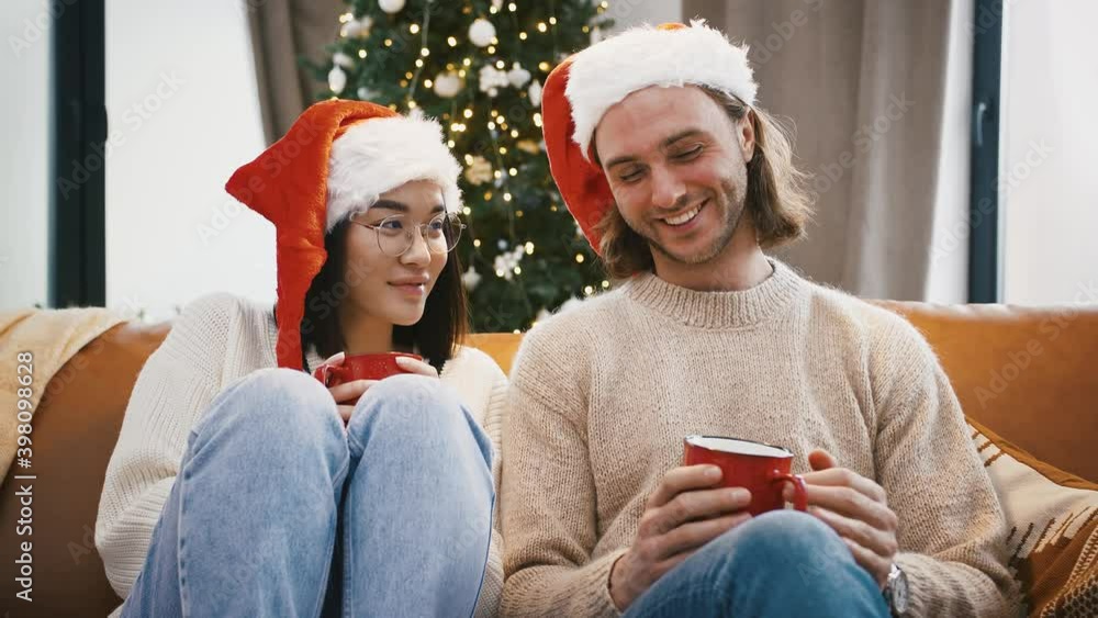 Multiracial male and female laughing, talking, holding mugs of tea, sitting on couch against decorated Christmas tree Multiracial male and female laughing, talking, holding mugs of tea, sitting on couch against decorated Christmas tree