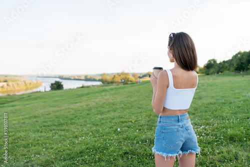 Wallpaper Mural Breakfast in nature, girl stands in park summer, looks river, free space for text, woman holds cup coffee in hands tea, weekend getaway in nature. Denim shorts, white tank top, green grass background. Torontodigital.ca