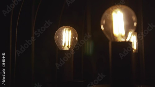 Transparent light bulbs in which the filament is visible are on and glow in a dark room on black metal rods stands
