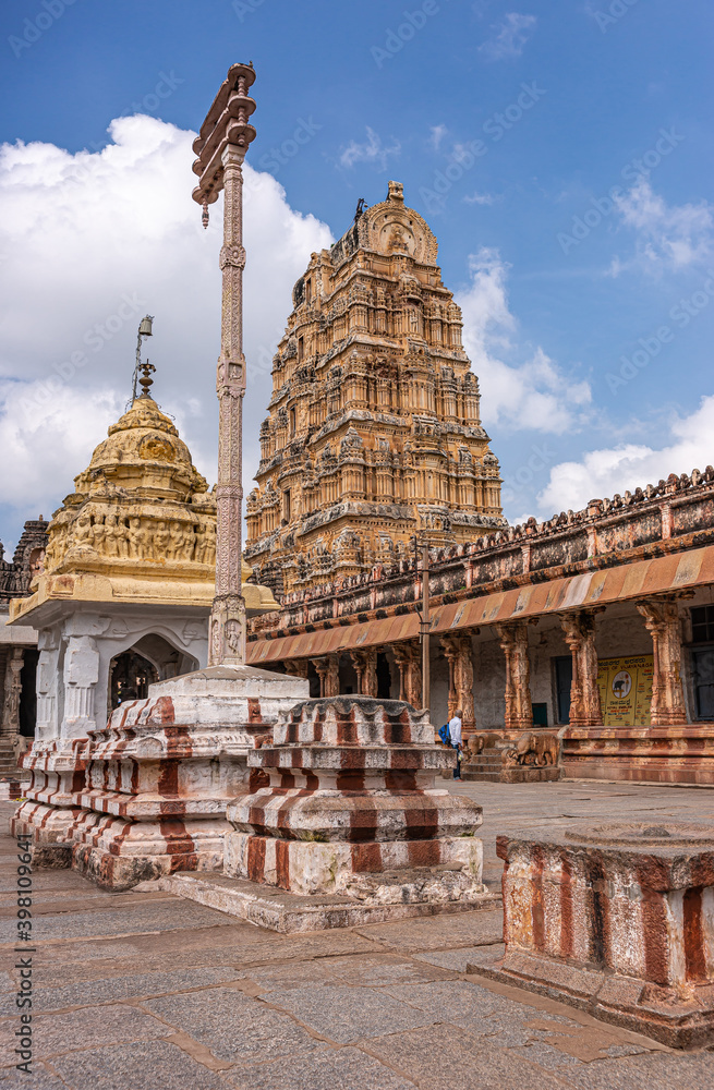 Hampi, Karnataka, India - November 4, 2013: Virupaksha Temple complex. Traditional Flagpost with yellow shrine, rusty pillars and brown stone of Shiva Vimanam under blue cloudscape.