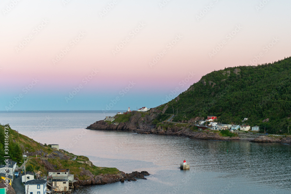 Sunrise over St. John's Harbour with some low lying clouds. The sky is pink near the horizon with calm water. There are mountains on both sides of the water. Houses are at the bases of both hills.