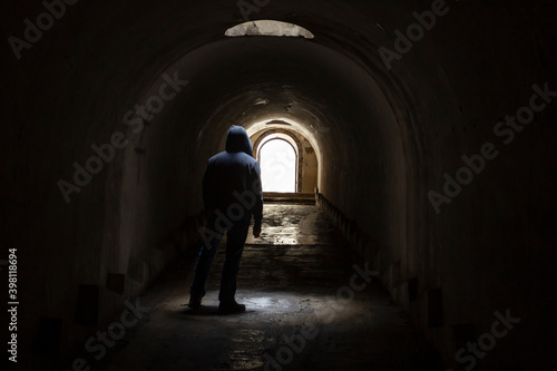 A hooded man walking towards a lighted exit from a dark  gloomy underground corridor. Light at the end of the tunnel leading to the exit to freedom.
