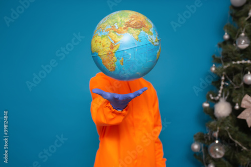Man in orange suit throws up globe while standing isolated on blue background near Christmas tree. Pandemic coronavirus 2019 concept.