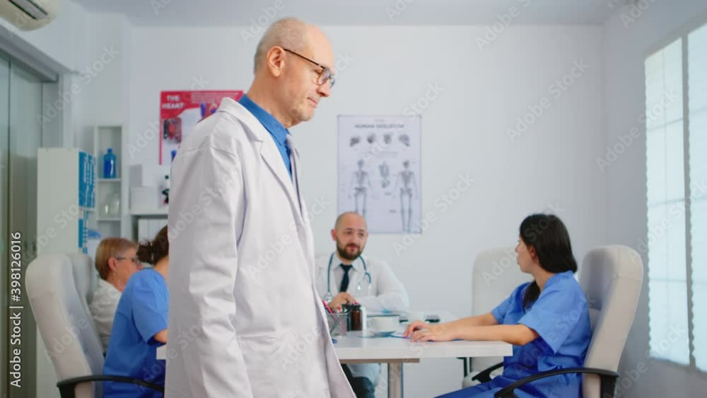 Portrait of elderly man surgeon standing in front of camera smiling in medical conference meeting office. Team of doctors working in background talking about symptoms of disease in clinic room Portrait of elderly man surgeon standing in front of camera smiling in medical conference meeting office. Team of doctors working in background talking about symptoms of disease in clinic room