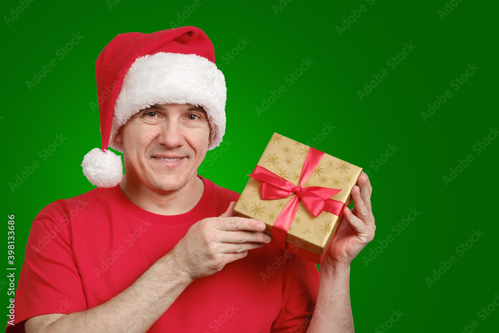 Portrait of a middle-aged smiling man in Santa hat showing gift box and looking at camera on green background Portrait of a middle-aged smiling man in Santa hat showing gift box and looking at camera on green background