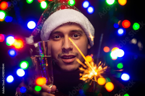 Young guy with a glass of champagne and a sparkler. Christmas garland. Blurred foreground. New Year s bokeh. Light of New Year s lights. Happy New Year.