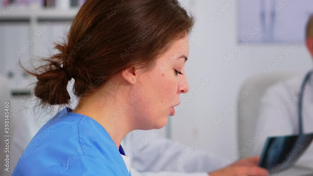 Close up of young woman nurse discussing with colleague during medical conference sitting on desk in hospital meeting office. Talking about symptoms of disease in clinic room during brainstorming Close up of young woman nurse discussing with colleague during medical conference sitting on desk in hospital meeting office. Talking about symptoms of disease in clinic room during brainstorming