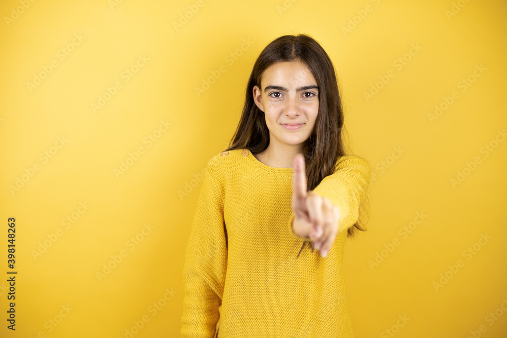 Pretty girl wearing a yellow sweater standing over isolated yellow background showing and pointing up with fingers number one while is serious Pretty girl wearing a yellow sweater standing over isolated yellow background showing and pointing up with fingers number one while is serious
