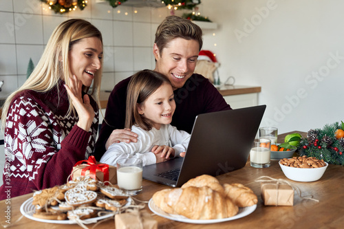 Happy family young parents with cute small kid child daughter waving hands using laptop computer at home sitting at Christmas table having virtual party dinner on video conference call together.