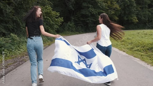 Two beautiful friends holding Israel national flag and going in a park. Independence Day.