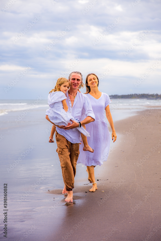 Happy family spending time at the beach. Summer holidays. Family vacation. Mother, father and daughter walking along the beach. Father carrying daughter in his arms. Copy space.