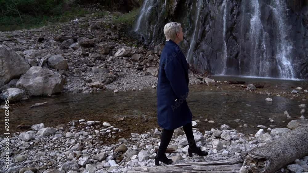 A middle-aged woman with short blond hair walks on a rocky surface near a waterfall and admires the calm nature on a cold day, sits on a log. She's wearing a blue coat. A middle-aged woman with short blond hair walks on a rocky surface near a waterfall and admires the calm nature on a cold day, sits on a log. She's wearing a blue coat.
