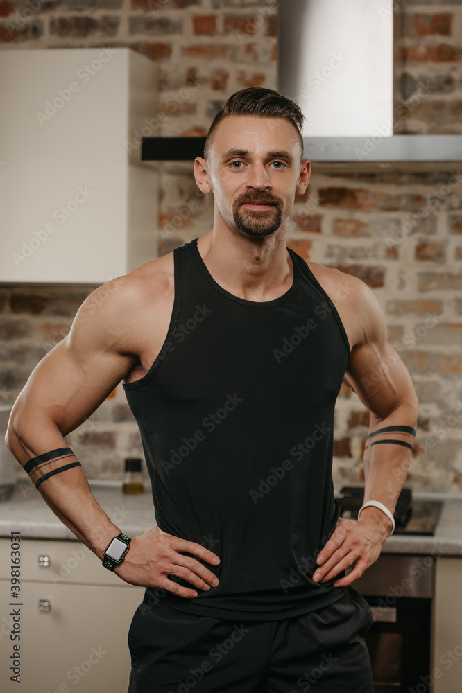 Obraz premium A muscular man with a beard is posing in his apartment after a workout. The athletic guy is demonstrating his sporty physique. A smartwatch and white rubber band are on the wrists.