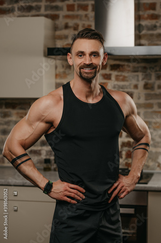 A smiling muscular man with a beard is posing with his hands on a waist in his apartment after a workout. An athletic guy with tattoos on his arms is demonstrating his sporty physique at home.