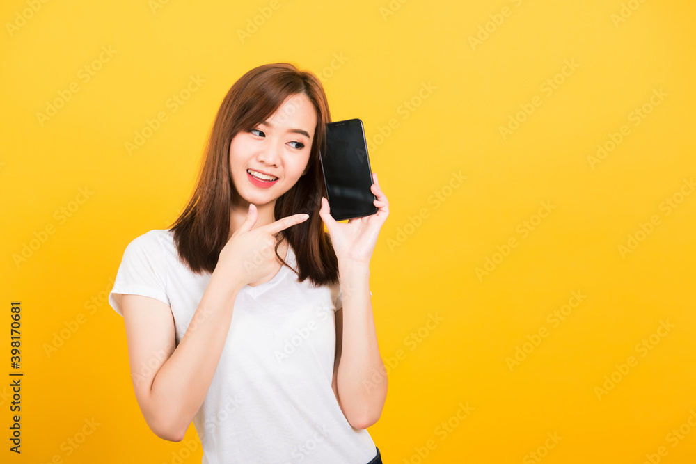 Asian happy portrait beautiful cute young woman smile standing wear t-shirt making finger pointing on smartphone blank screen looking to screen isolated, studio shot yellow background with copy space Asian happy portrait beautiful cute young woman smile standing wear t-shirt making finger pointing on smartphone blank screen looking to screen isolated, studio shot yellow background with copy space