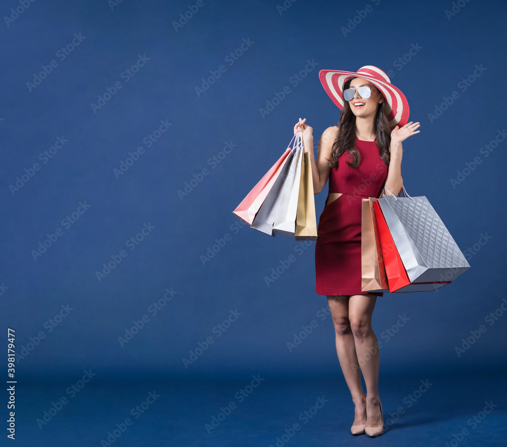 Happy young Asian woman wearing sunglasses and carrying multi color shopping bags on blue color background, exciting and looking at sales discount, black friday and consumerism concept, summer fashion