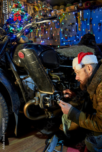 A man in a santa claus hat repairs a motorcycle in the garage on New Year's Eve.
