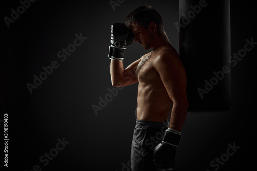 muscular handsome male boxer in black gloves standing near punching bag on dark background. copy space for text