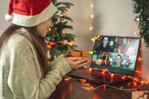 Woman in a red Santa hat is congratulating Christmas to friends or family remotely and holding a gift box in her hands
