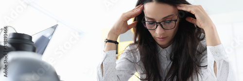 Woman sits with her eyes closed and holds her head in office. Overstrain at work and reporting concept photo