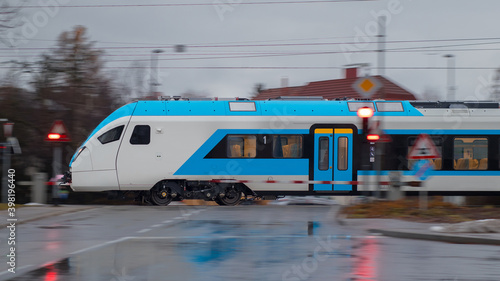 Side view of modern electric passenger train during cold winter rain is rushing through a grade crossing with road with barriers down and red signals flashing