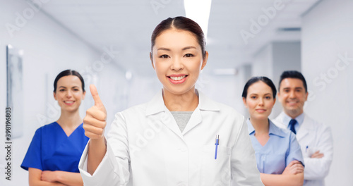 medicine, profession and healthcare concept - happy smiling asian female doctor in white coat showing thumbs up with medical team at hospital on background