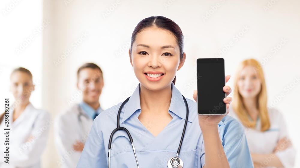 medicine, profession and healthcare concept - happy smiling asian female doctor or nurse in blue uniform with stethoscope showing smartphone with medical team at hospital on background medicine, profession and healthcare concept - happy smiling asian female doctor or nurse in blue uniform with stethoscope showing smartphone with medical team at hospital on background