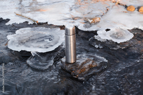 Thermos standing in a winter stream. Vacuum steel flask for hiking. Travel concept, warming drink in winter.