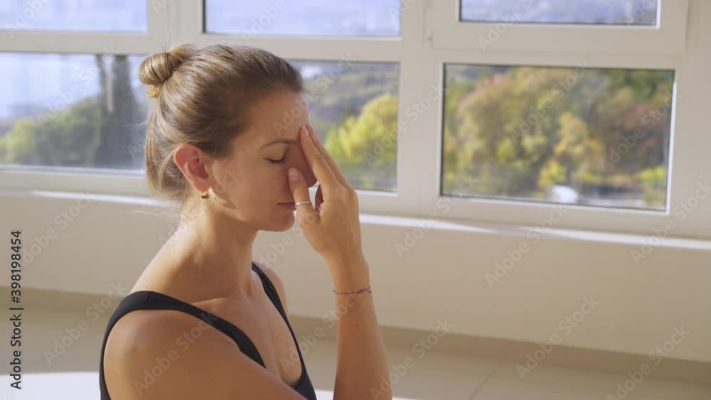 Pretty concentrated woman in black top does breathing gymnastic closing naris by finger near panoramic window in sunny gym at yoga training closeup Pretty concentrated woman in black top does breathing gymnastic closing naris by finger near panoramic window in sunny gym at yoga training closeup