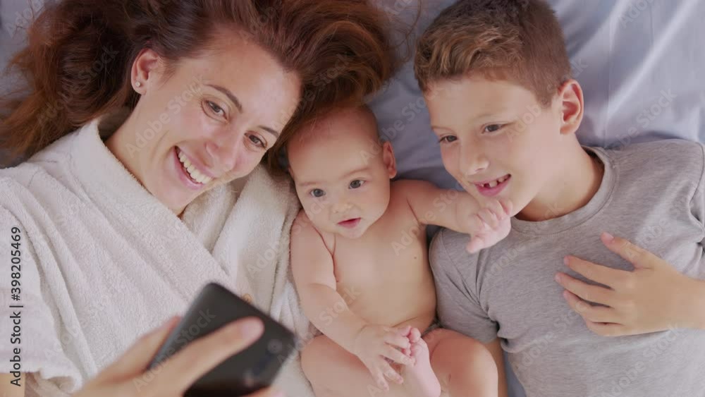 Happy mother with her kids are making a selfie or video call to father or relatives in a bed. Concept of technology, new generation,family, connection, parenthood, authenticity. Happy mother with her kids are making a selfie or video call to father or relatives in a bed. Concept of technology, new generation,family, connection, parenthood, authenticity.