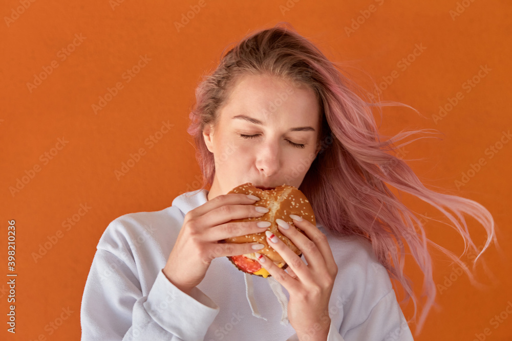 Beautiful cute young woman with pink hair and eating a Burger. The concept of harmful nutrition. Orange background. Beautiful cute young woman with pink hair and eating a Burger. The concept of harmful nutrition. Orange background.
