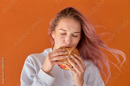 Beautiful cute young woman with pink hair and eating a Burger. The concept of harmful nutrition. Orange background.