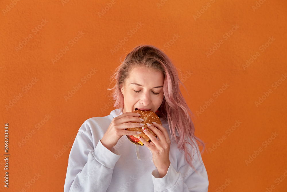 A woman on a diet eats a big Burger with an appetite. The concept of harmful nutrition. Orange background.