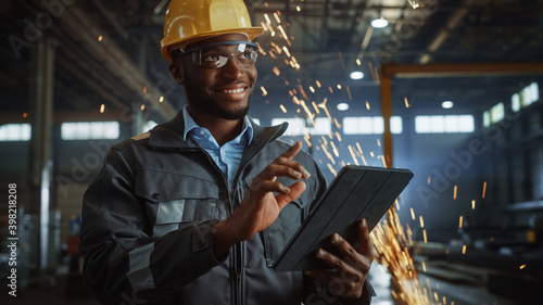 Professional Heavy Industry Engineer/Worker Wearing Safety Uniform and Hard Hat Uses Tablet Computer. Smiling African American Industrial Specialist Standing in a Metal Construction Manufacture.