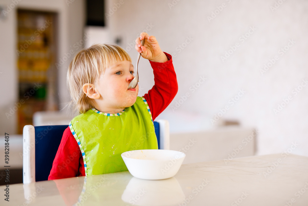 Cute baby 1,4 years old sitting on high children chair and eating vegetable alone in white kitchen Cute baby 1,4 years old sitting on high children chair and eating vegetable alone in white kitchen