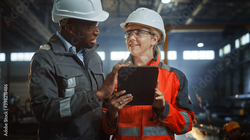 Two Heavy Industry Engineers Stand in Steel Metal Manufacturing Factory, Use Digital Tablet Computer and Have a Discussion. Black African American Industrial Specialist Talk to Female Technician. photo