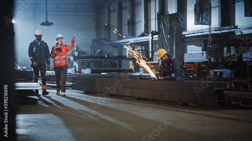 Two Heavy Industry Engineers Walk in Steel Factory, Use Tablet and Discuss Work. Industrial Worker Uses Angle Grinder in the Background. Black African American Specialist Talks to Female Technician. © Gorodenkoff