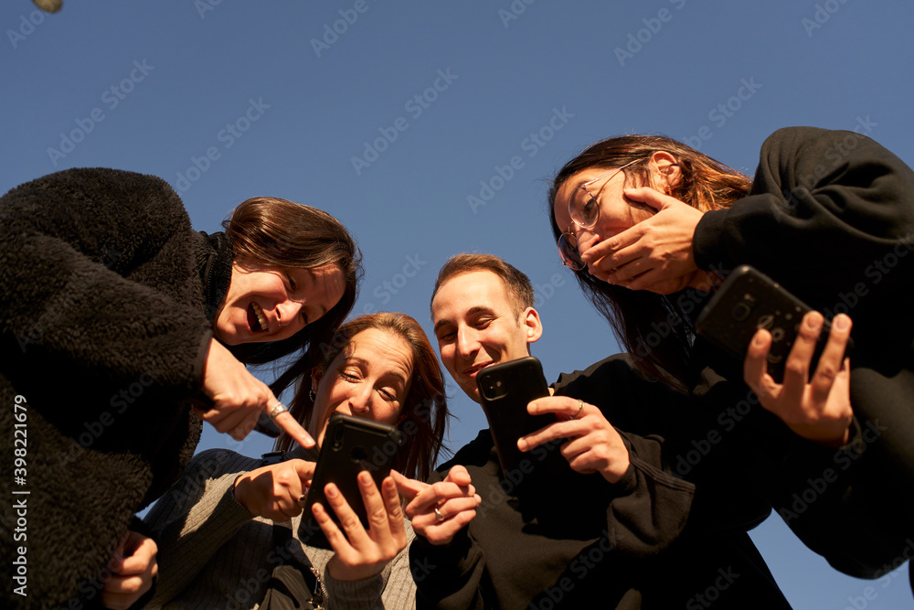 Obraz premium Low-angle of s group of friends using a smartphone and and laughing. He teaches them something fun on his phone. New normal lifestyle in the covid-19 season.