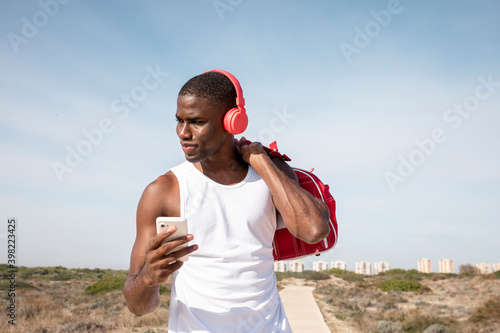 Vertical shot of young black athlete smiles while listening to music on his mobile with red headphones. Young man laughing while chatting.
