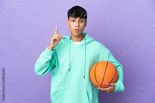 Young man playing basketball over isolated purple background intending to realizes the solution while lifting a finger up © luismolinero