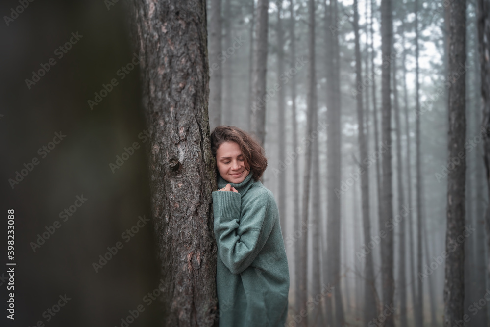 Happy woman in warm cozy green sweater in a beautiful foggy pine forest leaning against a tree trunk Happy woman in warm cozy green sweater in a beautiful foggy pine forest leaning against a tree trunk