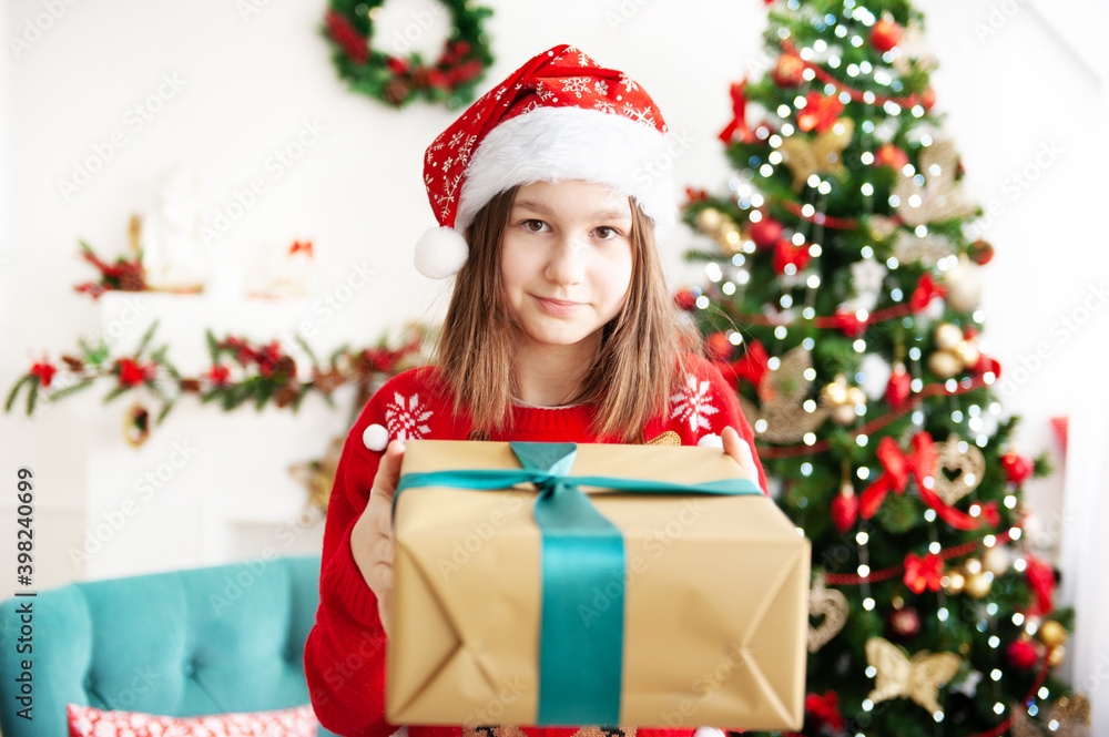 A young teenage girl in a Christmas cap and a red sweater with a large gift is sitting in a cozy chair next to the Christmas tree. Congratulation A young teenage girl in a Christmas cap and a red sweater with a large gift is sitting in a cozy chair next to the Christmas tree. Congratulation