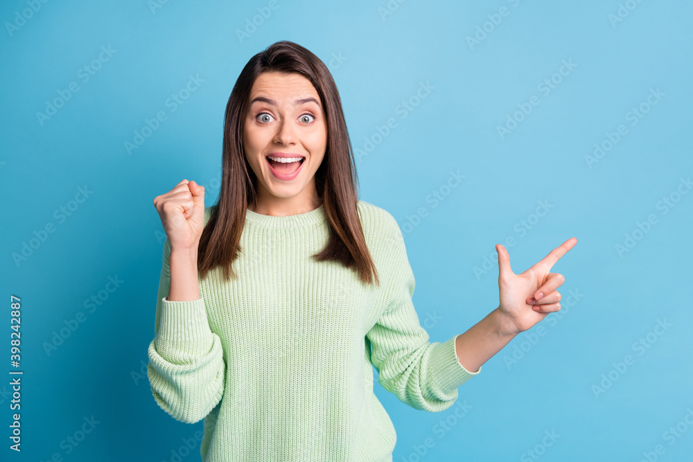 Photo of cute young lady indicate finger empty space raise fist open mouth wear green pullover isolated blue color background Photo of cute young lady indicate finger empty space raise fist open mouth wear green pullover isolated blue color background