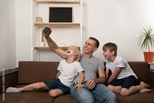 Young father and two sons take pictures of themselves on smartphone on the couch at home. Funny time