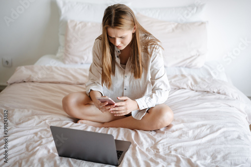 Beautiful serious nice girl using laptop and cellphone in bed at home