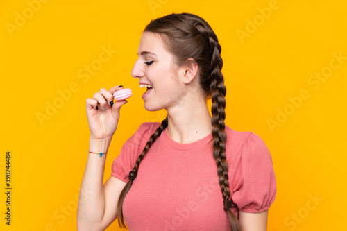 Young woman over isolated yellow background holding colorful French macarons and eating it