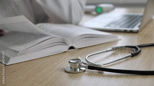 Work space of a doctor who reads books of medicine and research additional information in internet on a blurred background. Focus on stethoscope