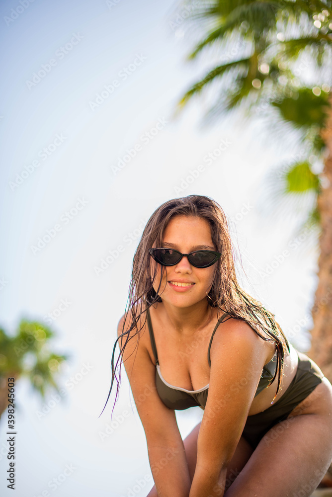 Fototapeta premium cute young teenage girl or woman posing and having fun on the beach under palm trees in summer swimsuit . Trendy girl posing. Funny and positive woman in sunglasses