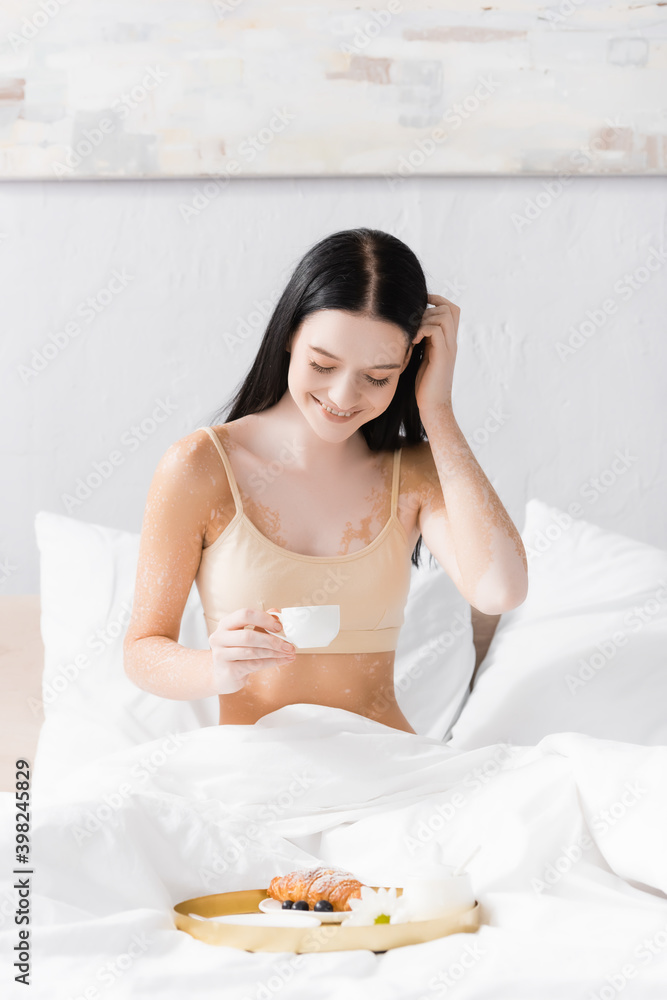 happy woman with vitiligo holding cup and fixing hair near breakfast tray happy woman with vitiligo holding cup and fixing hair near breakfast tray