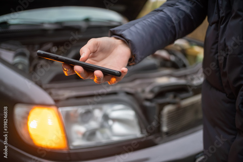 Woman's hand in a jacket holds a black mobile phone close-up against the open hood of a car on an emergency stop